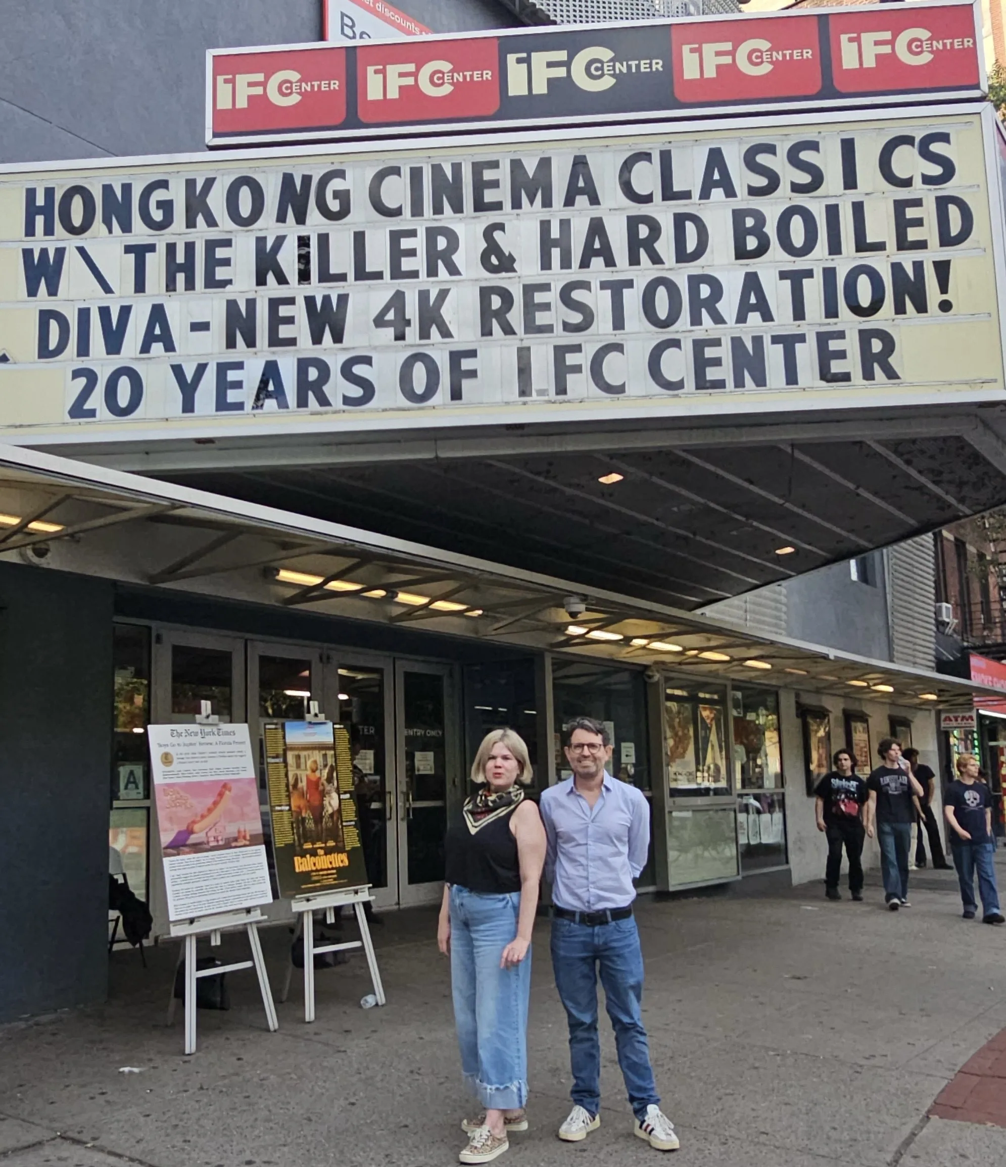 Two people stand in front of the IFC Center, a movie theater in the Greenwich Village neighborhood of Manhattan. At left is Caitlin Crowley, director of programs and promotions, a blond woman wearing pink sneakers, a sleeveless black top, and loose-fitting jeans with six-inch cuffs. At right is the Senior VP and General Manager of IFC Center, a slender white man with black-rimmed glasses, short brown hair flecked with gray, white sneakers with black stripes, dark blue jeans with a black belt, and a pale blue long-sleeved shirt. The marquee advertises two current offerings, a 4K restoration of Diva (1984) and a double feature of John Woo's The Killer (1989) and Hard-Boiled (1993).