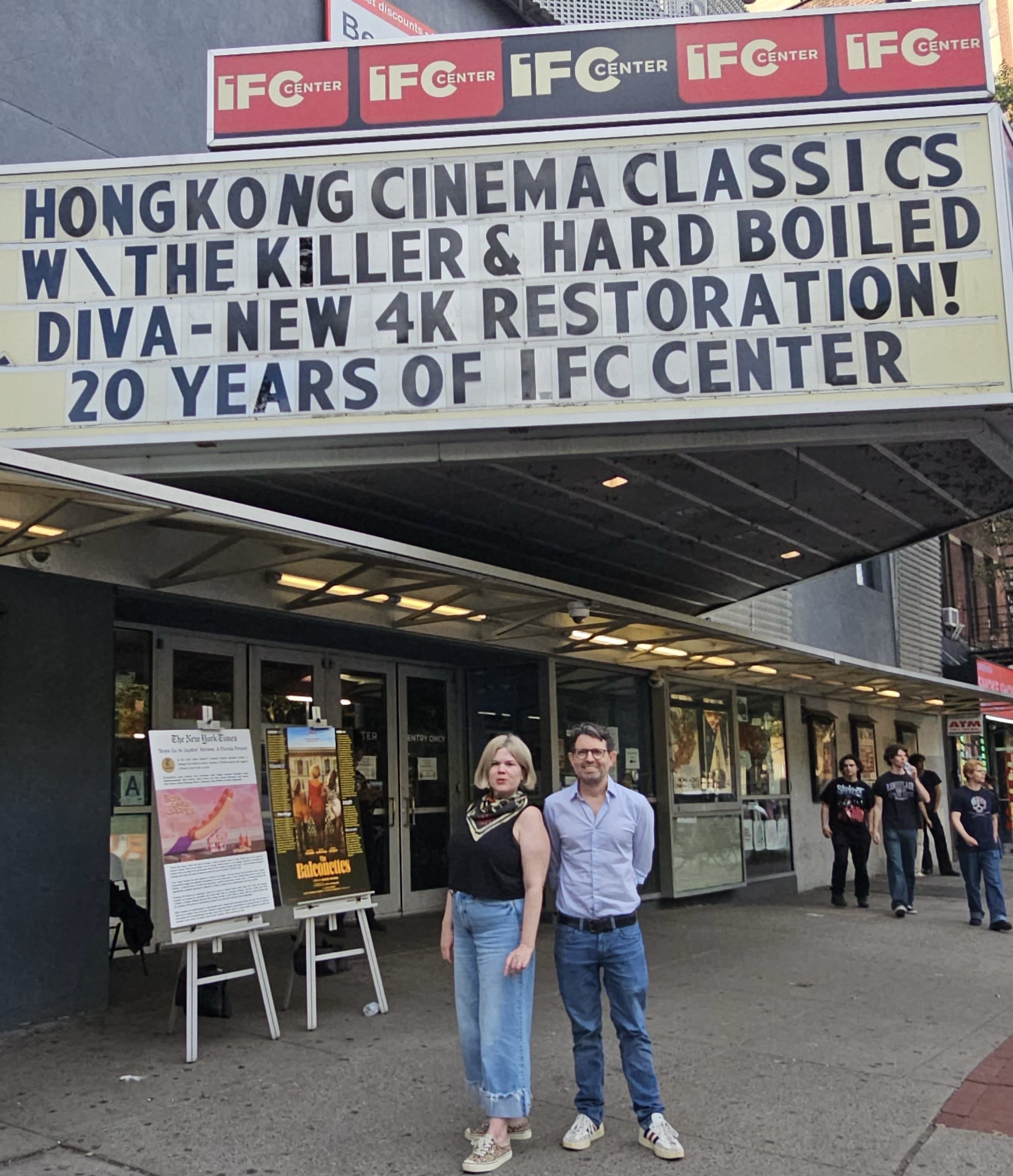 Two people stand in front of the IFC Center, a movie theater in the Greenwich Village neighborhood of Manhattan. At left is Caitlin Crowley, director of programs and promotions, a blond woman wearing pink sneakers, a sleeveless black top, and loose-fitting jeans with six-inch cuffs. At right is the Senior VP and General Manager of IFC Center, a slender white man with black-rimmed glasses, short brown hair flecked with gray, white sneakers with black stripes, dark blue jeans with a black belt, and a pale blue long-sleeved shirt. The marquee advertises two current offerings, a 4K restoration of Diva (1984) and a double feature of John Woo's The Killer (1989) and Hard-Boiled (1993).