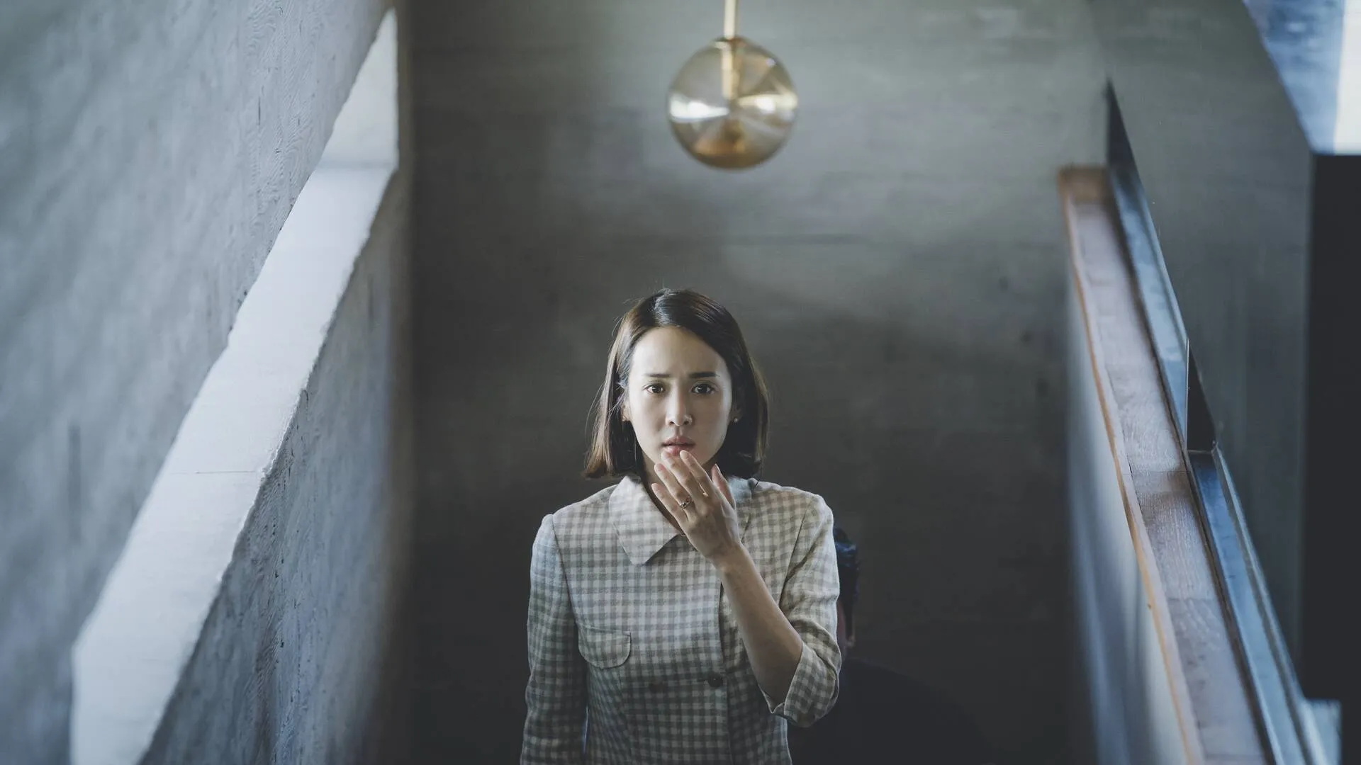 Cho Yeo-jeong in the climactic sequence of Bong Joon-Ho's 2019 thriller "Parasite" -- a finely featured Korean woman with a wide-collared blouse with an alternating pattern of grey and white plaid. She is raising her left hand to her chin after being shocked by what she just saw.