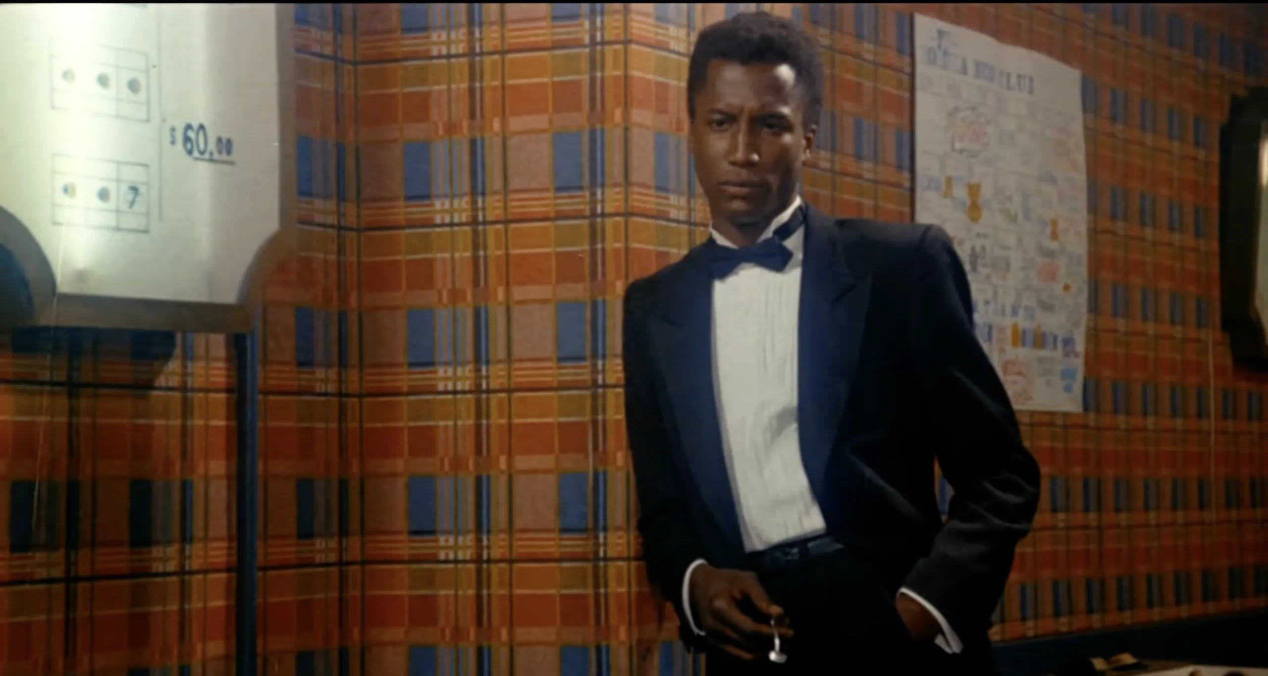 Michael Wright from a scene in the 1987 movie "Bedtime Eyes." He is standing against a wall wearing a black and white tuxedo.